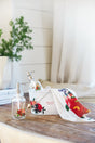A wooden table holds two glass soap dispensers with floral designs, a white container labeled Grove, and a white towel with red and yellow floral prints. A green potted plant sits in the background near a window.