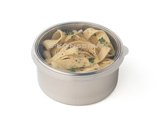A stainless steel container filled with wide pasta noodles garnished with herbs and ground black pepper, topped with a clear plastic lid.