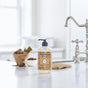 A bottle of Mrs. Meyers Hand Soap sits on a white kitchen countertop near a chrome faucet, with a small towel and a wooden bowl of fruit in the background.