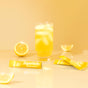 A glass of yellow drink with ice sits beside lemon halves and packets of SuperBelly Hydration Powder on a glossy, light yellow surface with a soft yellow background.
