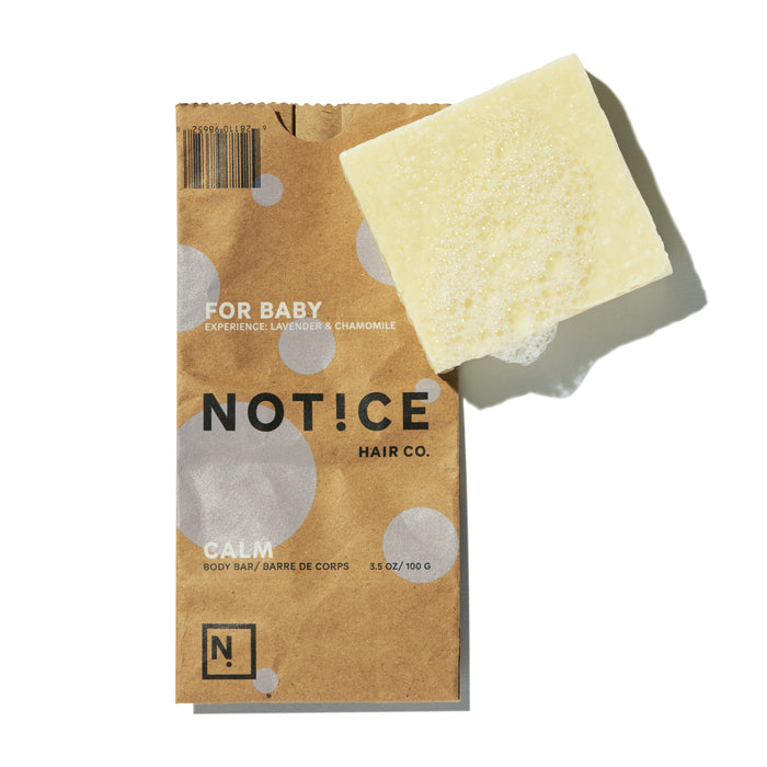 A rectangular beige bar of soap is partially resting on brown packaging labeled FOR BABY, EXPERIENCE: LAVENDER & CHAMOMILE, and NOTICE HAIR CO. CALM BODY BAR. The packaging has white polka dots.