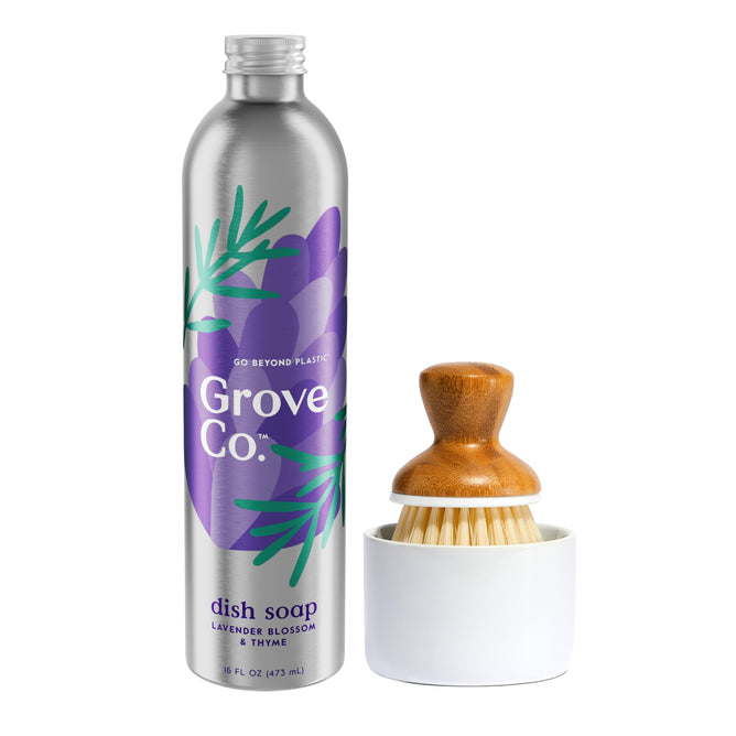 The Grove Co. Dish Washing Starter Set includes a tall silver bottle of Ultimate Dish Soap scented with lavender blossom and thyme, featuring botanical graphics to reduce plastic waste, along with a wooden-handled dish brush with bristles, resting in a white holder.