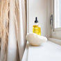 A glass dropper bottle of yellow facial oil labeled Terra is placed in a decorative white shell on a windowsill, with soft curtains and dried pampas grass in the background.