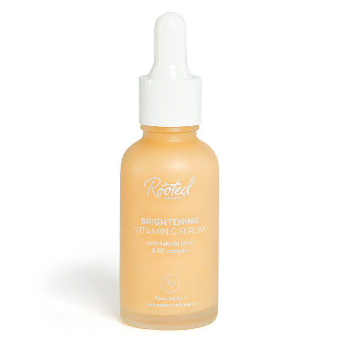 A frosted glass bottle with a white dropper cap labeled “Rooted Beauty Brightening Vitamin C Serum with kakadu plum & R7 complex.” The bottle contains light orange serum.