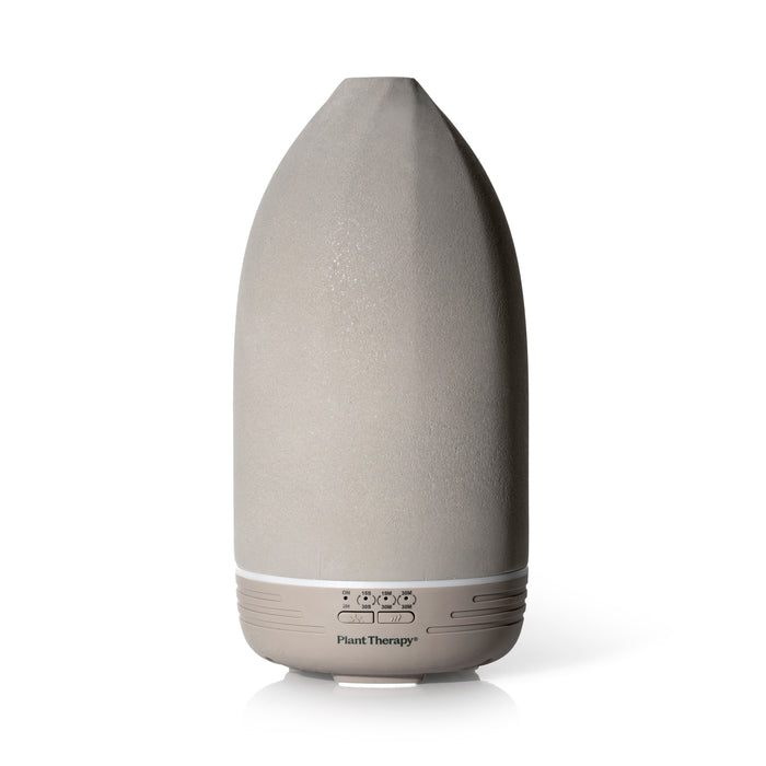 The Metro Stone Diffuser by Plant Therapy is a sleek gray ceramic device with a pointed top and white base, featuring control buttons. It elegantly stands on a white surface, ready to infuse your space with essential oils.