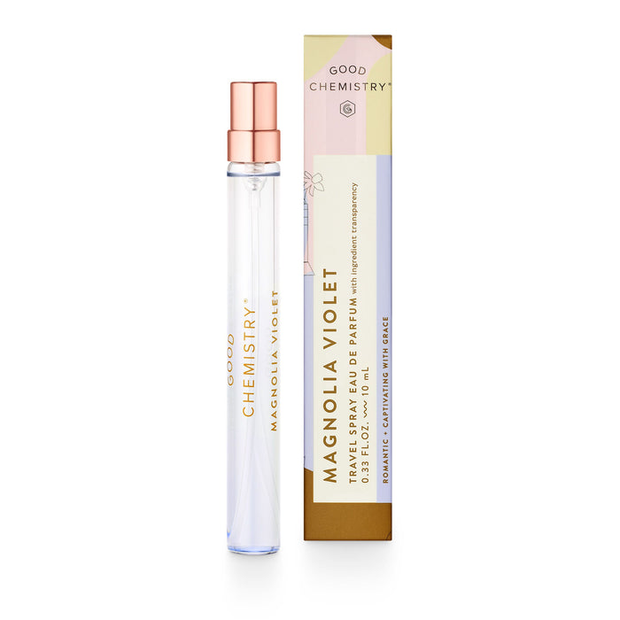 A clear glass perfume spray bottle labeled Good Chemistry Magnolia Violet stands next to its matching rectangular box decorated in white, peach, and gold tones. The box has floral accents and text describing the fragrance.