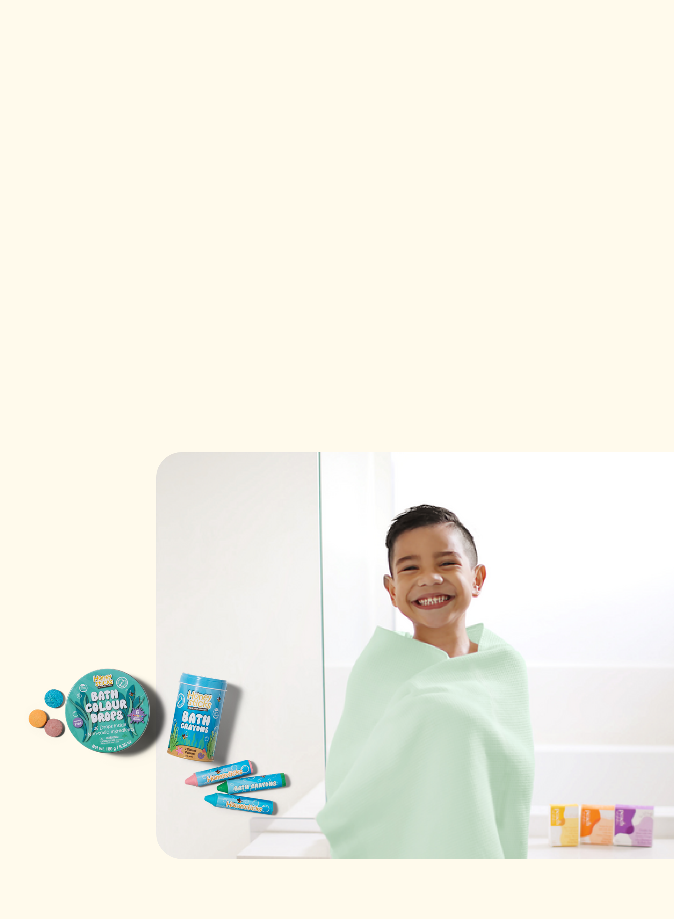 A smiling child wrapped in a light green towel stands by a bathtub. Bath crayons, a bath bomb, and colorful soap bars are displayed nearby on the left side of the image.