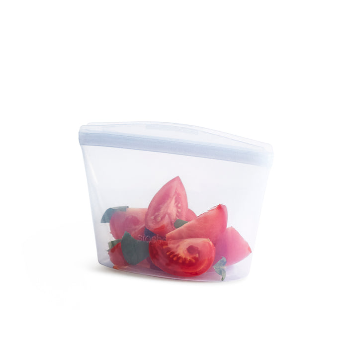 A clear reusable silicone bag containing sliced tomatoes and green leafy herbs, set against a white background.