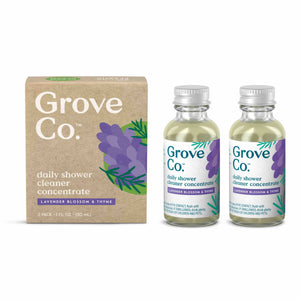 Two bottles of Grove Co. Daily Shower Cleaner Concentrate, featuring a plant-based formula with a lavender blossom and thyme scent, are displayed next to matching cardboard packaging adorned with purple lavender illustrations. Each bottle, designed to reduce plastic waste, includes a silver cap.