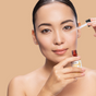 With hair pulled back, a person applies Tree To Tubs Skin Brightening Triple Action Vitamin C Serum for Face using a dropper. The soft beige background emphasizes the skincare routine.