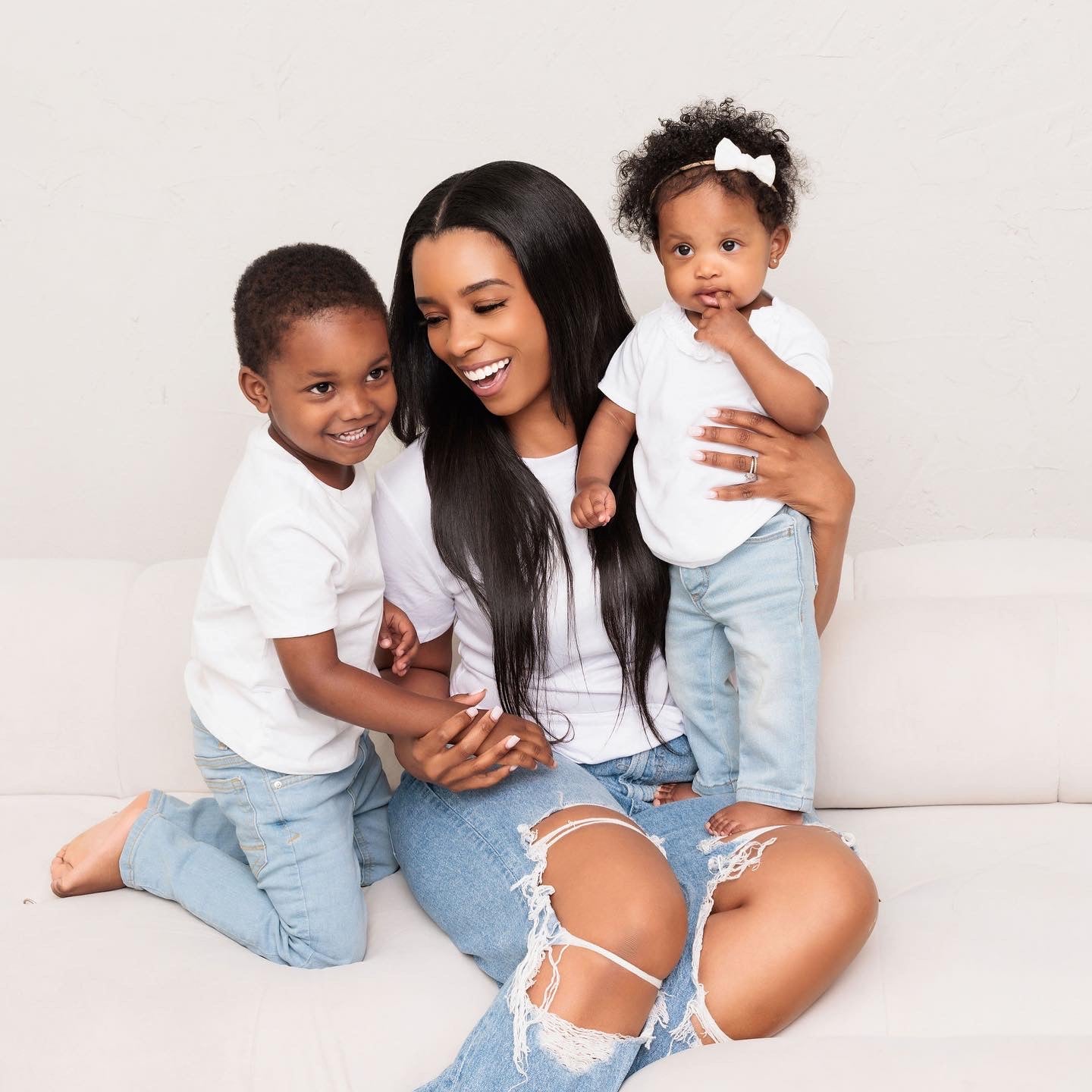 A woman sits on a white couch, smiling and holding a baby girl on her knee, while a young boy kneels beside her. All three are wearing white shirts and blue jeans. The baby has a white bow in her curly hair.