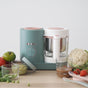 A baby food maker with a teal and pink design sits on a counter surrounded by fresh vegetables and glass containers of pureed baby food.