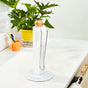 A roll of paper towels on a white holder with a wooden handle and metal arm, placed on a kitchen counter. In the background, there is a green potted plant and colorful cups near the sink.