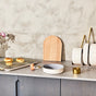 A modern kitchen countertop with a cooling rack holding four pastries, a wooden cutting board, a round dish, a gold measuring cup, and a white plate rack holding stacked white plates with gold-handled cutlery.