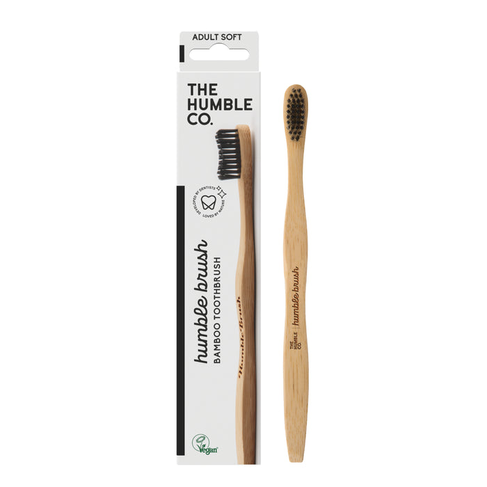 The Humble Co.s Adult Bamboo Toothbrush - Soft, featuring dentist-approved black nylon bristles, rests beside its vegan packaging. The white box has Humble Brush Bamboo Toothbrush in black text and is labeled Adult Soft.