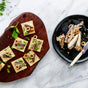 A wooden board holds six crackers topped with Patagonia Provisions White Anchovies Spicy, sliced green olives, and chopped herbs—each offering 22g protein. Nearby, a black plate displays more anchovies, olives, and a knife on marble.