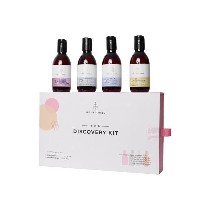 Four bottles of Holy Curls hair products stand on top of a white box labeled “The Discovery Kit.” The box lists contents and features simple, minimalist design elements.