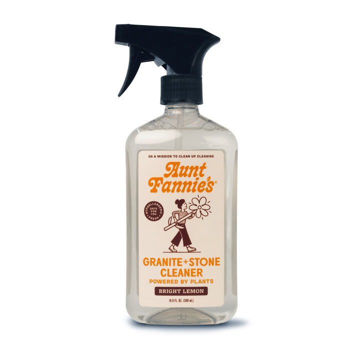 A clear spray bottle of Aunt Fannies Granite + Stone Cleaner, Bright Lemon scent, featuring a black trigger nozzle and a label with an illustration of a woman holding a flower. The label highlights its powered by plants.
