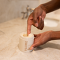 A person dipping their finger into a glycerin-rich container of PAUMEs Overnight Hand and Foot Hydration Mask on a marble bathroom countertop.
