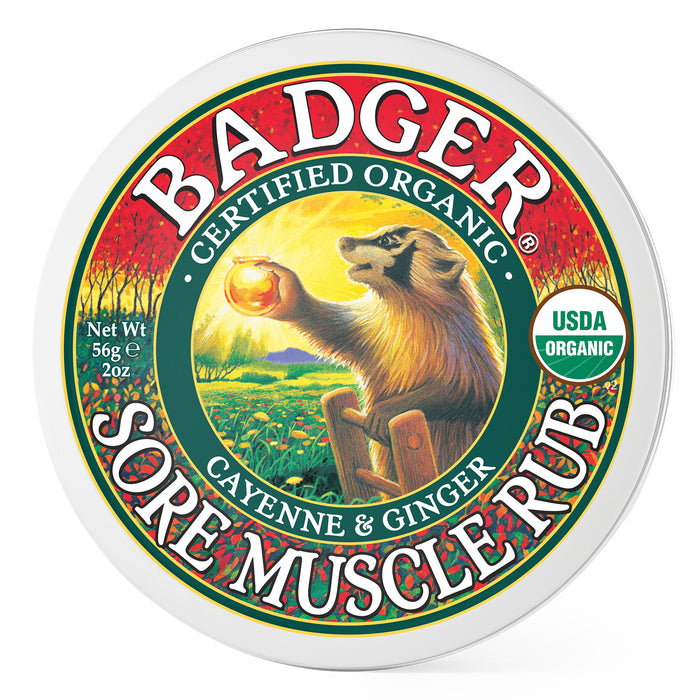 Round tin labeled Badger Sore Muscle Rub, Certified Organic, Cayenne & Ginger. Illustration of a badger holding a glowing orb, with a field and sunrise in the background. USDA Organic seal on right side. Net weight 2 oz.