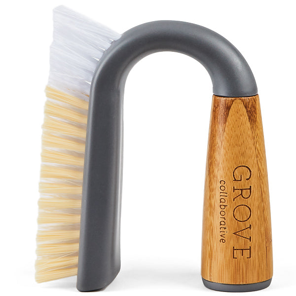 A dish brush with a curved gray handle, white and beige bristles, and a wooden grip engraved with the words “GROVE collaborative.”.