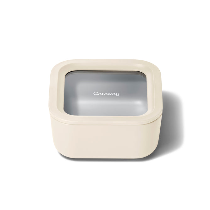 A cream-colored, square food storage container with a transparent lid displaying the brand name Caraway in white text. The container is shown on a white background with a soft shadow.