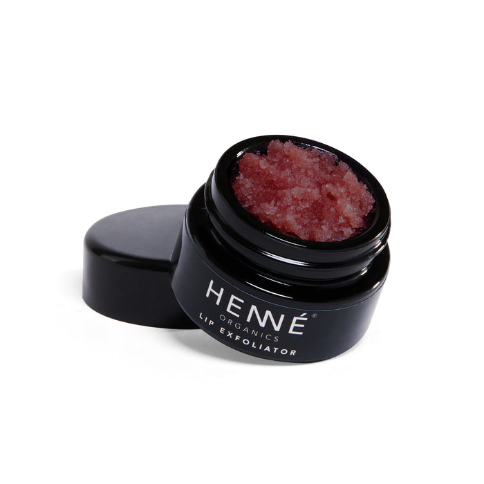 A small open black jar labeled “HENNÉ ORGANICS LIP EXFOLIATOR” is filled with a reddish, grainy scrub. The lid is resting beside the jar. The background is white.