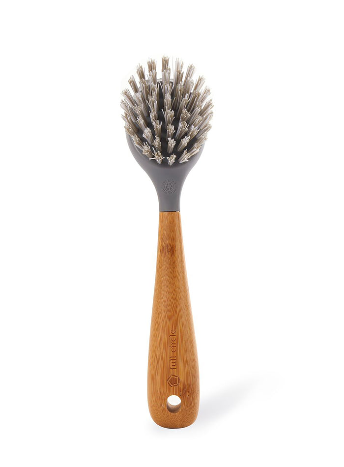 A dish brush with a round head, stiff bristles, a gray plastic top, and a wooden handle with a hole near the base for hanging. The background is plain white.