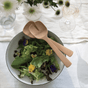 A fresh salad with leafy greens and edible flowers sits in a white bowl, accompanied by sleek Bambus Short Servers. The bowl rests on a white tablecloth with a glass of water, wine, and a vase of white flowers nearby, embodying modern design elegance.