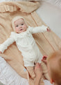 A baby in a white zip-up onesie and bonnet lies on a butter cream Little Love Baby Blanket by AU Baby while an adult’s hands adjust the fabric.