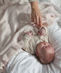 A baby lies on a white bed being tucked in with a soft, light pink AU Baby Little Love Blanket - Cloud, an organic cashmere baby blanket that features a ruffled edge.