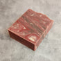 A rectangular bar of marbled soap featuring swirling patterns of red, pink, white, and gray, placed on a light gray textured surface.