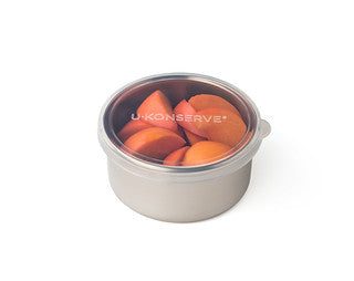 A round stainless steel container with a clear plastic lid, holding several orange fruit slices, shown on a white background. The lid is labeled U-Konserve.