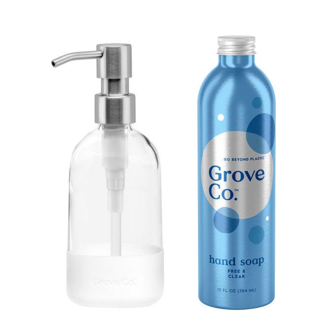 A clear glass soap dispenser with a sleek silver pump stands beside a blue metal bottle from the Grove Co. Hand Soap Starter Set, highlighting circles and text that emphasize the brands commitment to reducing plastic waste with its eco-friendly hand soap refill option.