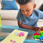 A young child sits on a couch, focusing as they arrange HABA’s Logic! GAMES: Happy Worms, made from sustainably sourced wood, to match shapes on a yellow activity card on the glass table in front of them.