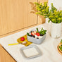 A bowl of yogurt topped with strawberries, blackberries, and mint sits on a counter next to a yellow napkin, a spoon, and a vase with yellow flowers. The bowl’s lid rests nearby on the marble surface.