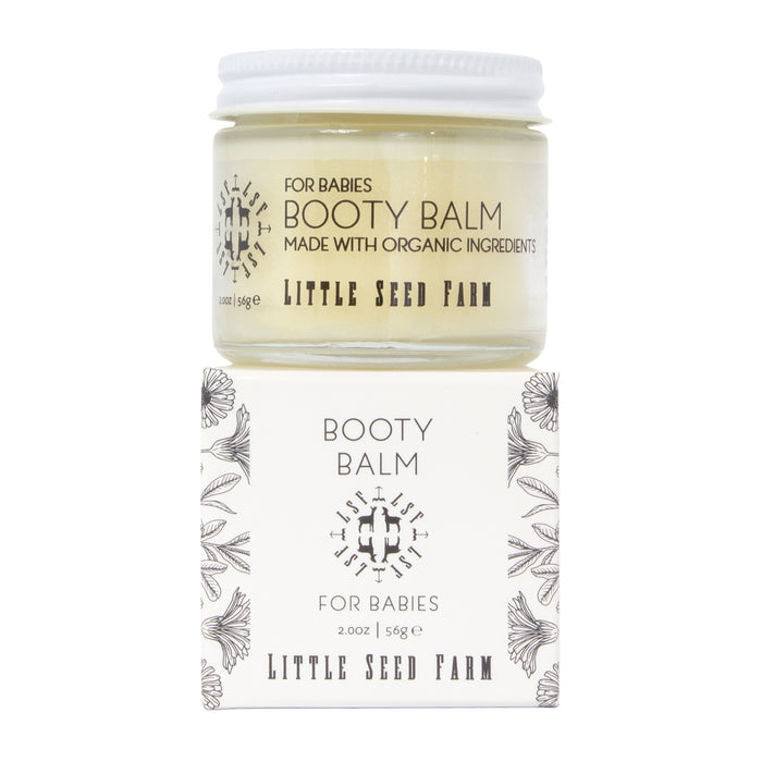 A jar of Little Seed Farm Booty Balm for Babies, a cloth diaper safe organic balm, rests on its matching box. The white packaging features black floral designs and text showcasing its gentle, healing herbs for babies.