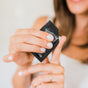 A woman in a white top with light nail polish squeezes Little Seed Farm Deodorant Cream Sample - Activated Charcoal from a small black packet onto her finger. Her face is partially visible.