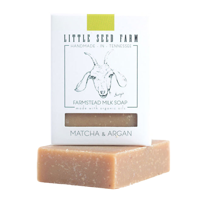 A Matcha & Argan Bar from Little Seed Farm, with a beige, slightly rough texture, rests partly out of its white box featuring the brand name and an illustrated goat.