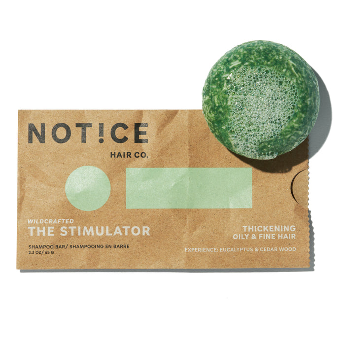 A green shampoo bar with bubbles rests on a brown paper package labeled NOT!CE Hair Co. The Stimulator for thickening oily & fine hair, with eucalyptus and cedar wood scent.