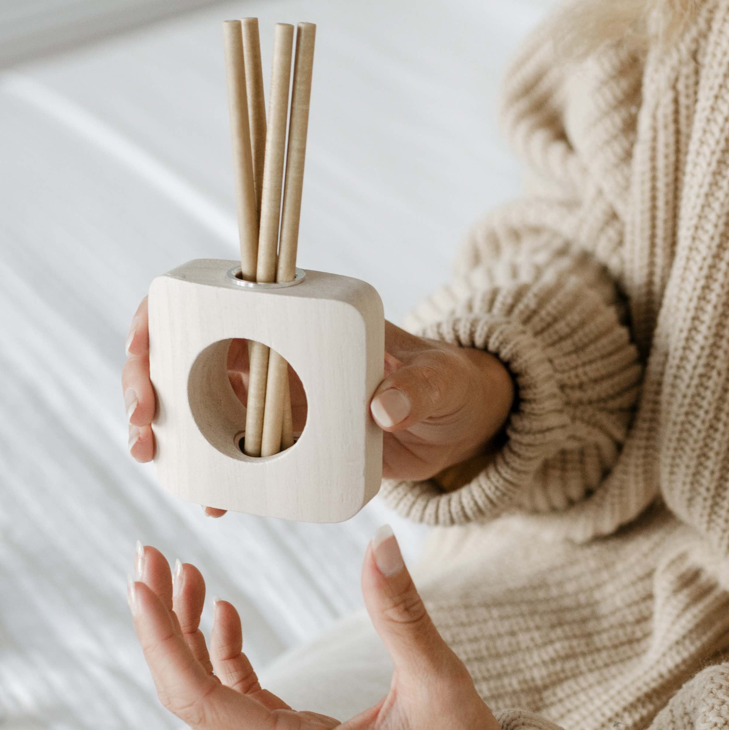 A person in a beige knit sweater holds a wooden diffuser with reed sticks, resting on their lap. The scene suggests a calm and cozy environment.