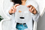 A person in a striped shirt sitting on a white wicker chair holds an ORGAID Vitamin C & Revitalizing Organic Sheet Mask made with Ecoderma fabric technology. Their face is partly hidden by the organic mask, and they have long, curly hair.