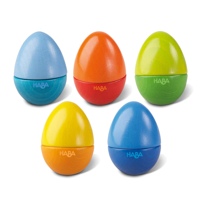 Five colorful egg-shaped wooden shakers by HABA are arranged in two rows. The eggs are blue, orange, green, yellow, and a darker blue, each with a matte finish and the brand name “HABA” on the bottom half.