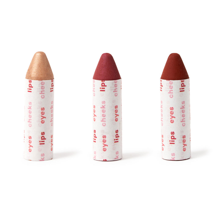 Three crayon-shaped makeup sticks in gold, pink, and red shades, each wrapped in white paper with red text reading lips, eyes, and cheeks, standing upright against a white background.