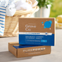 Close-up of two Grove Co. PurePower Enzymatic Laundry Detergent Sheets boxes, featuring 35 Loads and 2-in-1 Enzyme Stain Fighting Power, highlighting reduced plastic waste. A softly blurred laundry basket with clothes appears in the background.
