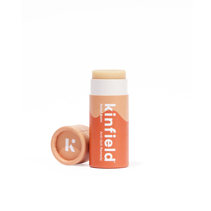 An orange tube of Kinfield Relief Balm, labeled Itch-Fix Remedy, stands upright with its cap, displaying a white K logo, lying on its side beside it. The balm is exposed, against a white background.