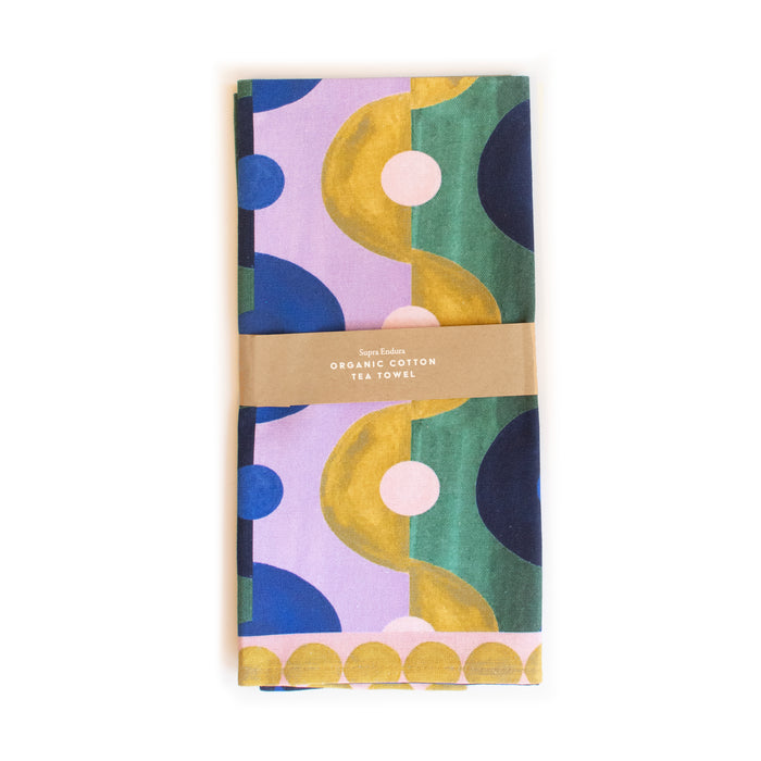 A folded tea towel with a bold, colorful abstract pattern in purple, green, yellow, and dark blue, wrapped with a brown paper band labeled Organic Cotton Tea Towel.