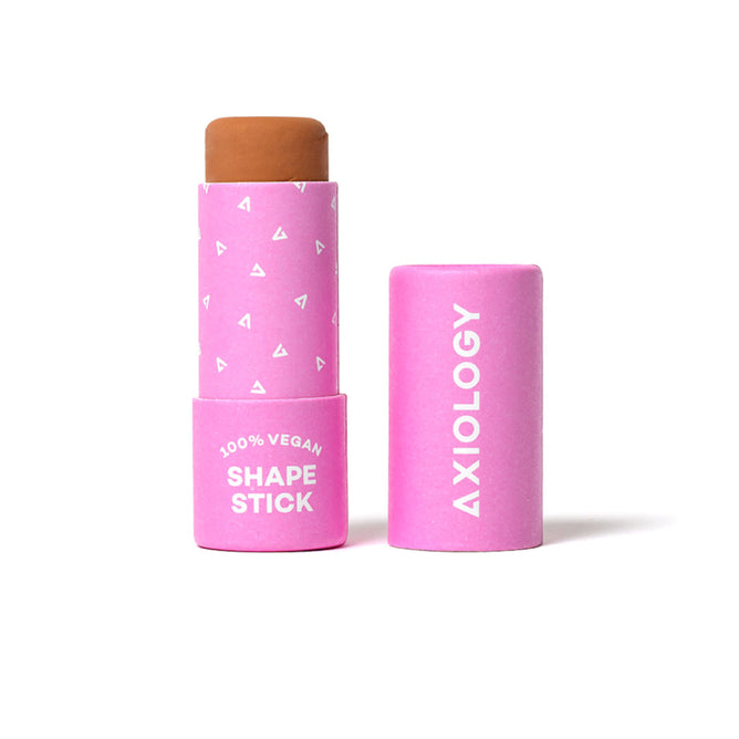 A brown makeup stick in a pink tube labeled 100% VEGAN SHAPE STICK with the brand name AXIOLOGY on the lid, which is placed next to the stick. Small white triangle patterns decorate the tube.