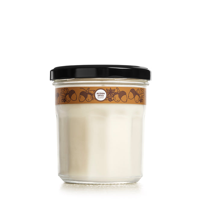 The glass jar features a black lid and a brown label with acorns, berries, and leaves illustrations, plus a circular logo. It holds Mrs. Meyers Acorn Spice Soy Candle.