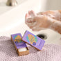 Two boxes of Grove Co. Hand Soap Sheets (40 Washes) sit on a lavender towel by the sink, with someone in the background creating suds under running water infused with a gentle, natural fragrance.